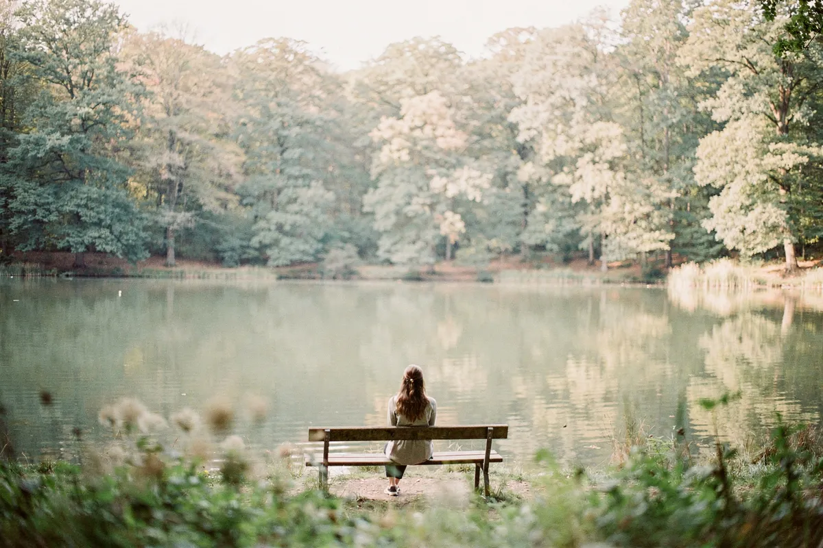 Une femme assise sur un banc en bois face à un lac entouré de grands arbres feuillus